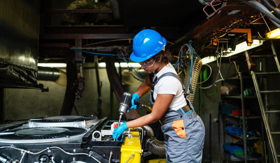 Young female technician wearing safety glasses and hardhat using a power drill to repair industrial machinery in a factory