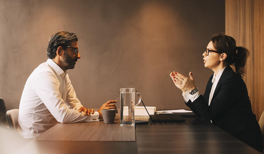 Two people sit across from each other at a conference table in a modern office setting, engaged in a professional discussion.