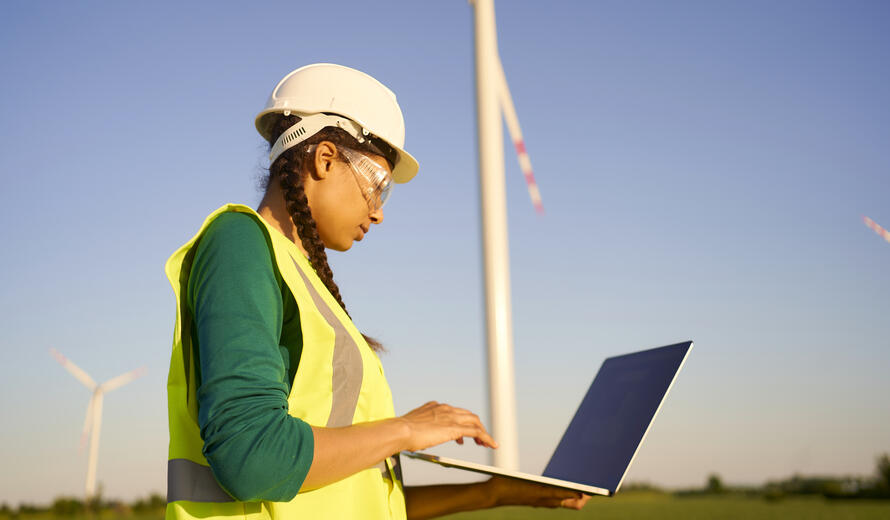 Female engineer wearing hard hat and reflective green jacket standing with laptop against wind turbine and setting it up. Energy concept.