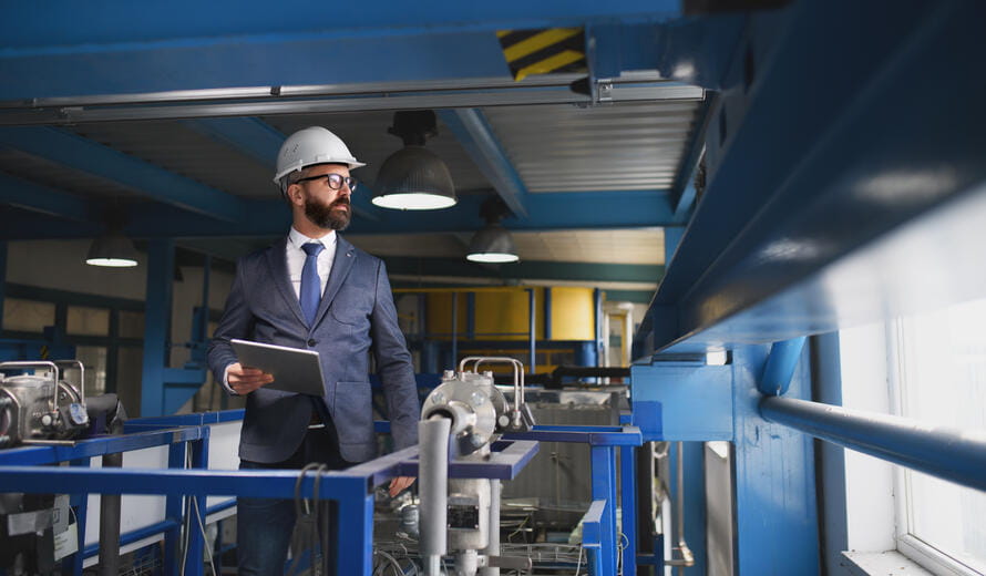 A chief Engineer in the hard hat walks through industrial factory while holding tablet.