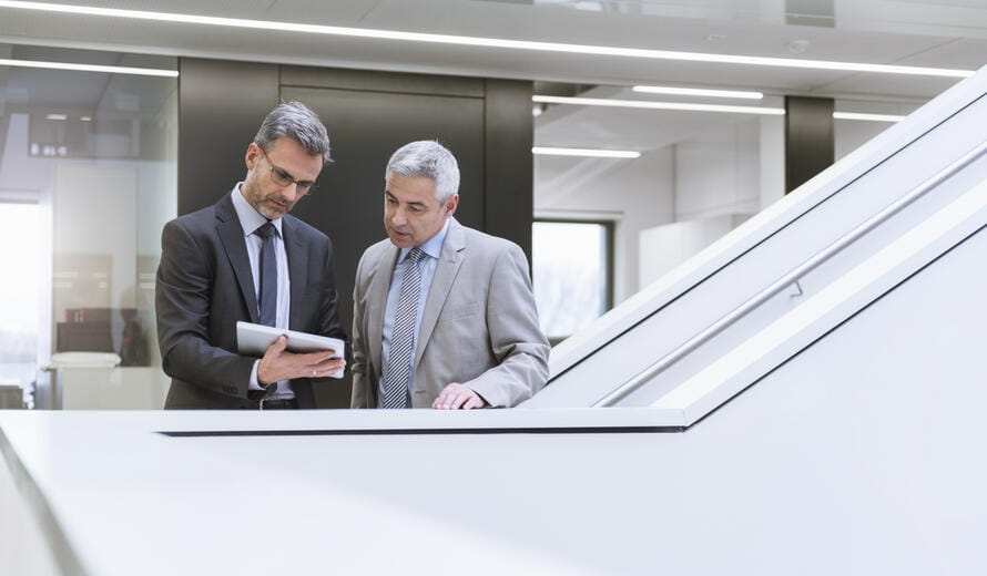 Two business professionals stand together in a modern office space, looking at a tablet and discussing information