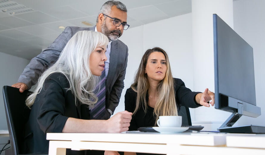 Business group watching presentation on pc monitor and discussing project, sitting at workplace with cup of coffee and pointing at display. Business communication concept