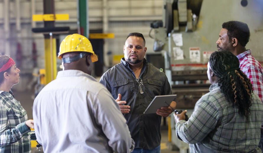 A group of five multiracial mature adults, two women and three men, working in a metal fabrication shop having a meeting on the factory floor. 