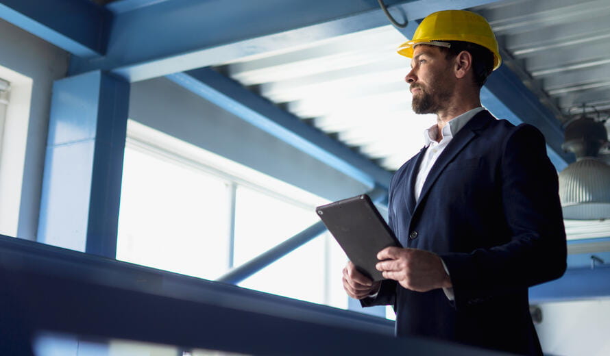 Portrait of businessman with protective helmet in warehouse, holding digital tablet. Low angle view.