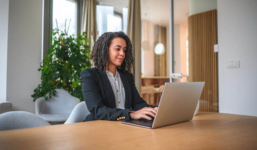 Mid adult Hispanic female working with a laptop in a corporate office. She is sitting at her desk, typing and sending emails.