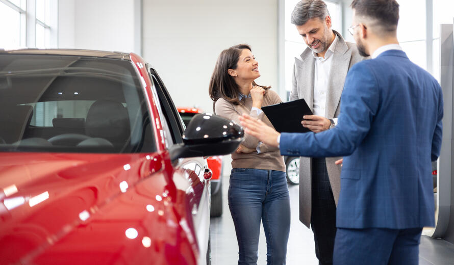 Salesman with a digital tablet showing vehicle features to an excited couple at a car dealership