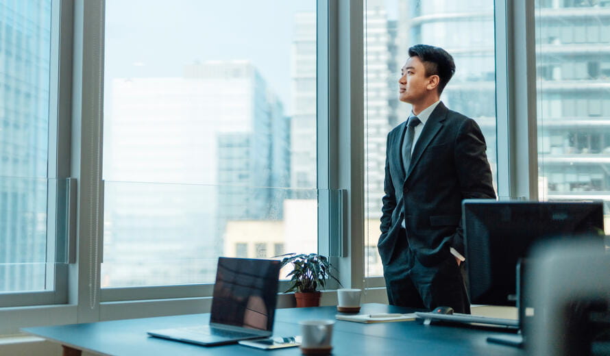 Thoughtful young Asian businessman looking out window, standing at desk in a bright and modern office.