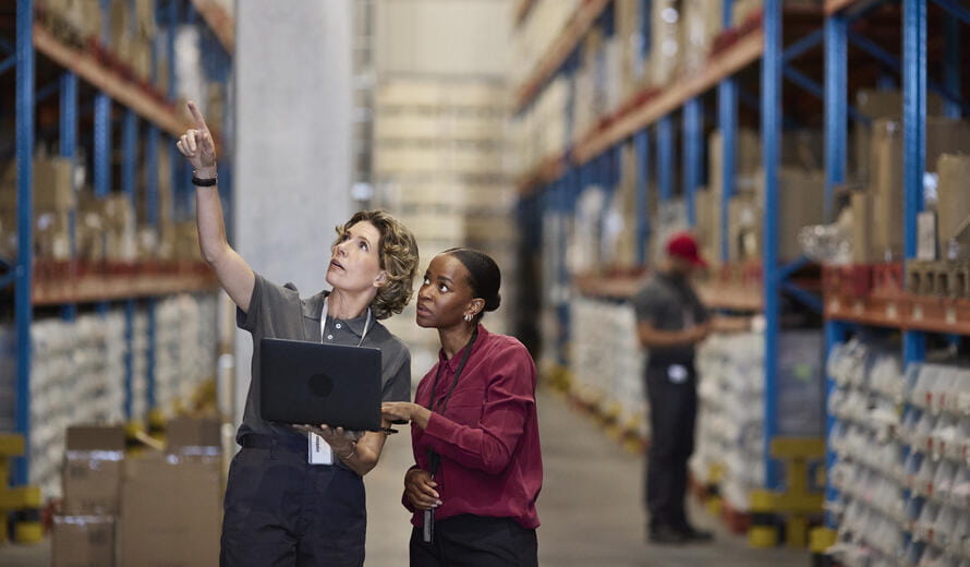 Mature female worker holding laptop and pointing while talking with manager in warehouse