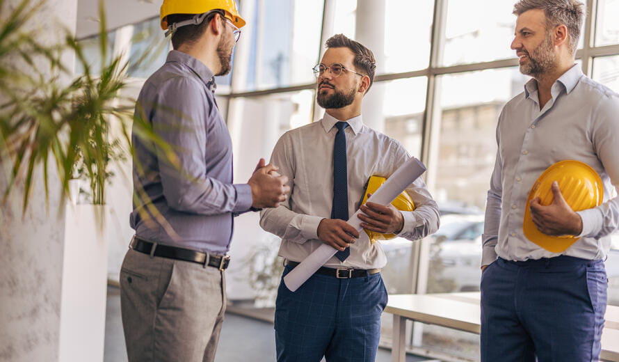 Three male engineers and architects discussing a new project in a modern office, collaborating over blueprints while wearing hardhats and dressed in formal attire