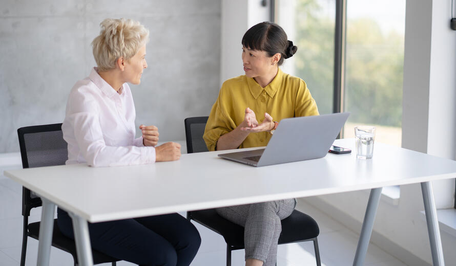 A productive exchange of ideas occurs as two women share insights using a laptop in a bright, open office environment.