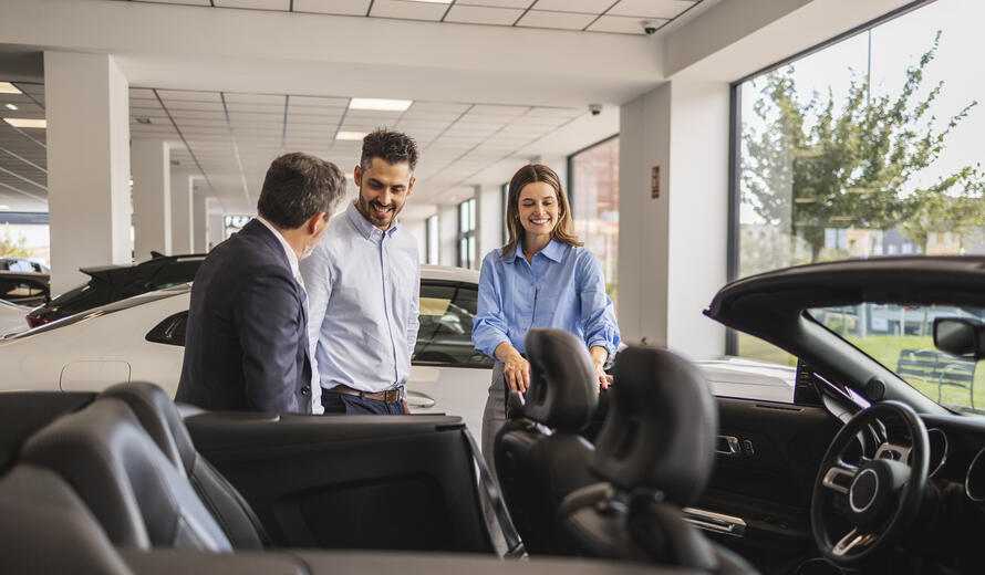 Happy couple admiring a new convertible car in an auto dealership showroom, engaging with a professional car seller who is assisting them with their purchase decision