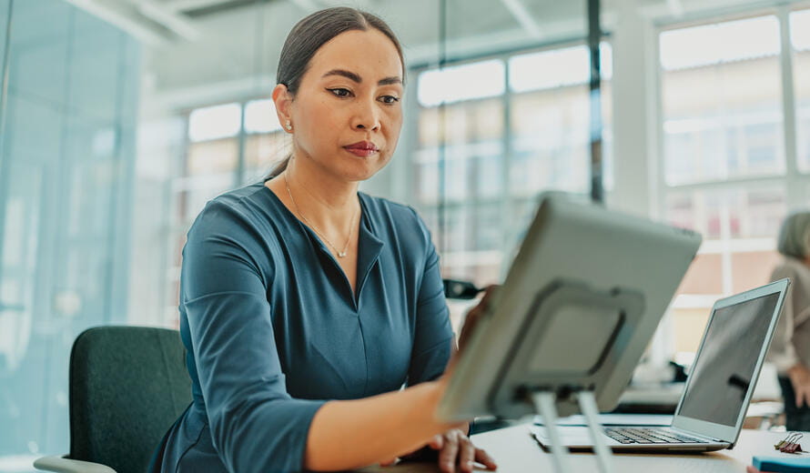 Stock image showing a professional seated at a desk in a modern office environment, using a tablet device alongside a laptop and desk accessories.