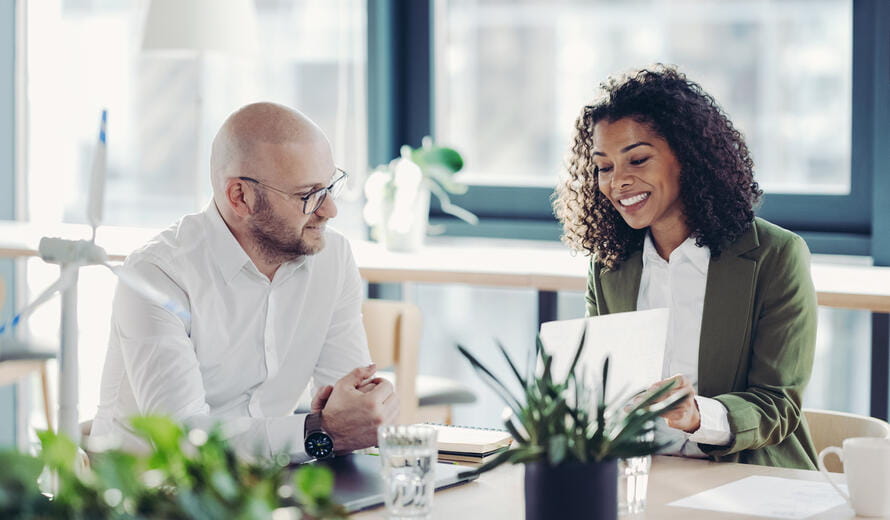 Two business professionals sitting together in a modern green office, reviewing a document and discussing work‑related matters.
