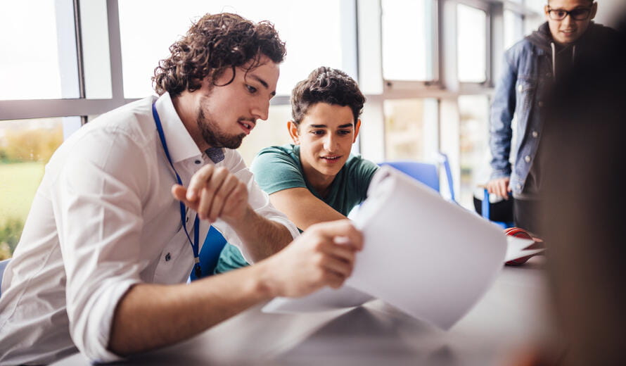 Teacher showing a student some results on his paperwork. The student looks happy as they sit side by side at a table. Another student looks on.