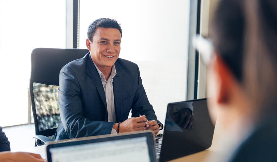 Stock-style professional image depicting a business meeting environment with participants engaged in discussion and laptop-based collaboration, suitable for use in consulting, advisory, or corporate strategy contexts.