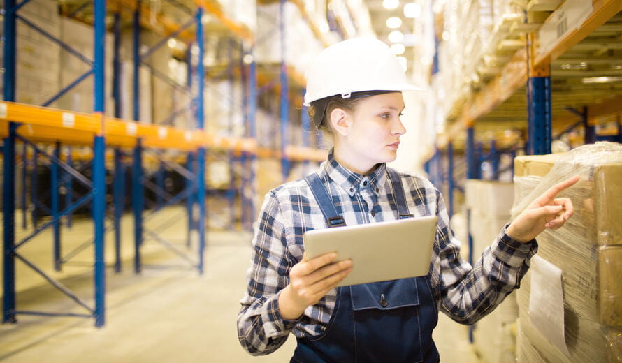 Serious busy young female supervising expert using tablet while examining count of packages in warehouse