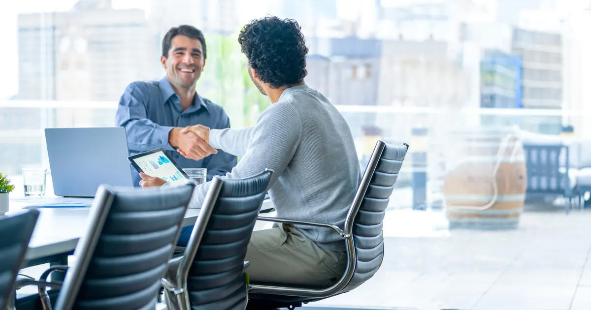 Two businessmen working on a digital tablet and laptop computer in the office in a board room or meeting room. They are shaking hands closing the deal and a large window behind them
